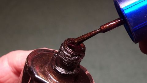 Ein Hand hält einen Nagellackpinsel über einer offenen Flasche mit hartnäckigem Glitzer-Nagellack in einem neutralen Raum.