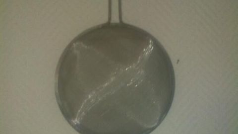 Ein rundes Küchensieb hängt an einer Wand; sein Metallgeflecht reflektiert das Licht schwach im Innenraum.