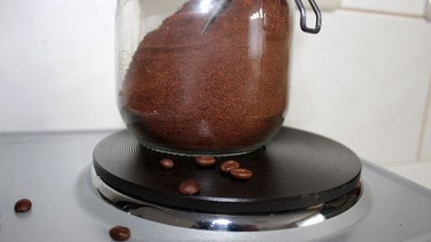 Eine schwarze Herdplatte hat Kaffeepulver neben einem Glasbehälter, der gefüllt ist, und einige Kaffeebohnen liegen verstreut.