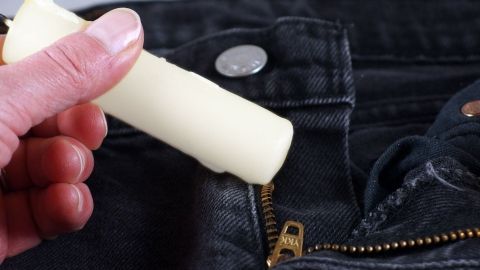 Eine Hand hält eine Kerze, die über einen klemmenden Reißverschluss einer Jeans gestrichen wird, um ihn zu lösen.