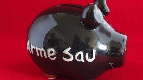 Ein schwarzes Sparschwein steht auf rotem Hintergrund und zeigt die Aufschrift „Arme Sau“. Es symbolisiert Geld sparen.