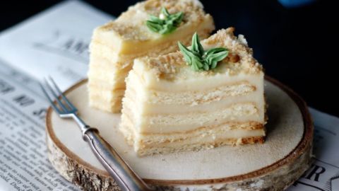 Zwei Stücke Buttercremekuchen liegen auf einer Holzplatte, umgeben von Zeitungsseiten, mit einer Gabel daneben.