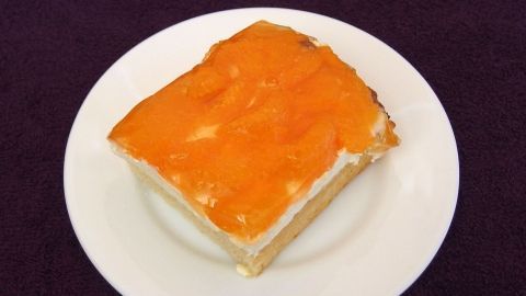 Ein Stück Schmandkuchen liegt auf einem weißen Teller, mit einer glänzenden orangefarbenen Oberfläche und einem geschichteten Geschmack.