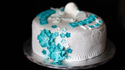 Ein dekorativer Kuchen mit weißen und blauen Zuckergussblumen steht auf einem transparenten Teller vor dunklem Hintergrund.