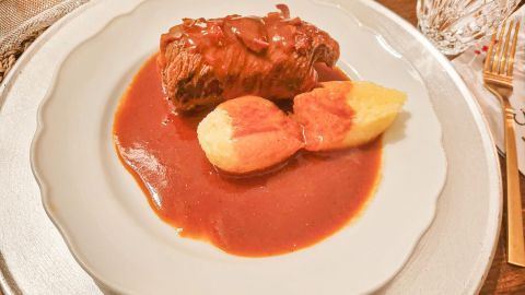 Eine gefüllte Rinderroulade liegt auf einem Teller, umgeben von einer würzigen Sauce und Beilagen wie Kartoffeln.