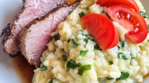 Das Risotto aus Fenchel liegt mit frischen Kräutern und Tomatenscheiben auf dem Teller, begleitet von zartem Fleisch.
