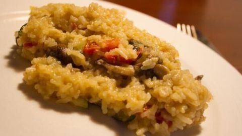 Ein Teller Risotto mit Tomaten und Pilzen liegt auf einem weißen Teller, auf einem Holztisch im warmen Licht.