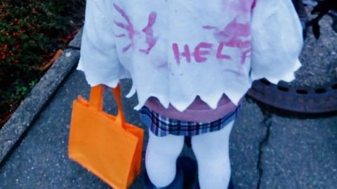 Ein kleines Mädchen trägt ein zerrissenes Kostüm mit der Aufschrift „HELP“ und hält eine orangefarbene Tüte.