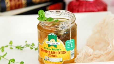 Du kannst die letzten Reste aus deinem Honigglas verwerten, indem du einen Teebeutel hineinlegst und ihn mit heißem Wasser übergießt. Ein Honigglas mit Sommerblütenhonig steht auf einem dekorativen Tablett, umgeben von frischen Minzblättern.