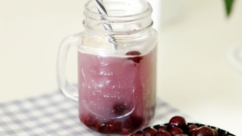 Eine Glasbecher mit Sauerkirschbowle steht auf einem karierten Untersetzer, umgeben von frischen Kirschfrüchten.