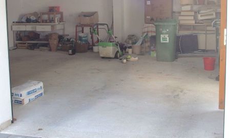 Der Raum zeigt eine Garage mit einem Ölfleck auf dem Betonboden. Im Hintergrund stehen diverse Gerätschaften und Kartons.