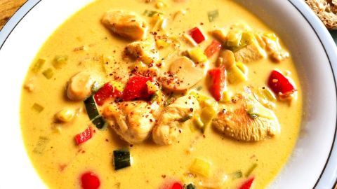 Die Gemüsesuppe in einer weißen Schüssel, cremig und bunt, wird mit frischem Gemüse und Hähnchenstücken serviert.