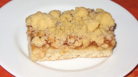 Ein Stück Apfel-Quark-Streuselkuchen aus Hefeteig liegt auf einem weißen Teller, umgeben von rotem Tischdeckenstoff.