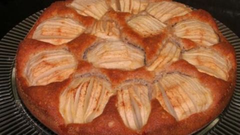 Eine Apfeltorte mit goldbrauner Kruste und applierten Äpfeln liegt auf einem gedeckten Tisch in einer dunklen Umgebung.