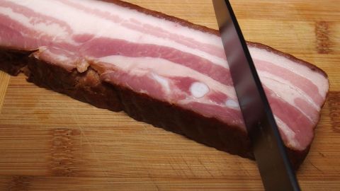 Ein dicker Streifen Speck liegt auf einem Holzbrett, während ein scharfer Messer einen Schnitt ansetzt, um ihn zu teilen.