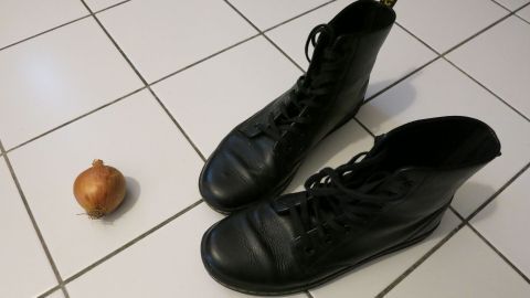 Durch Abreiben mit einer Zwiebel verschwinden Wasserränder an Schuhen: Keine Angst, die Zwiebel macht keine anderen Flecken! Schwarze Lederschuhe stehen auf einem Fliesenboden neben einer rohen Zwiebel, die zur Reinigung der Schuhe genutzt wird.