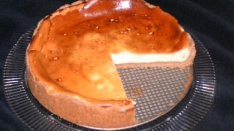 Käsekuchen mit einem Stück herausgeschnitten ruht auf einem gläsernen Tortenteller, platziert auf dunklem Stoff.