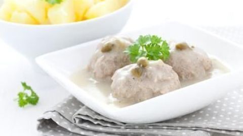Drei saftige Königsberger Klopse liegen in einer cremigen Soße, begleitet von Kartoffeln in einer Schüssel im Hintergrund.