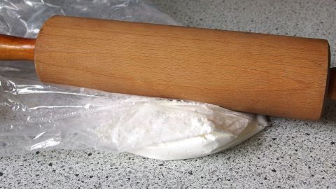 Hart gewordenen Puderzucker kann man mit dem Nudelholz wieder fein machen. Ein Nudelholz liegt auf einem Haufen Puderzucker, bereit zum Ausrollen auf einer grauen Arbeitsfläche.