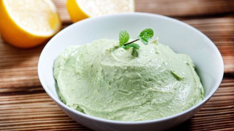 Eine cremige Avocado-Dip-Variation befindet sich in einer weißen Schüssel, umgeben von frischen Zitronenhälften.