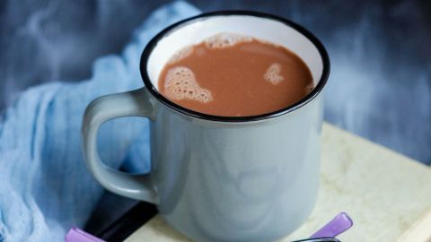 Ein grauer Becher mit heißen Schokoladenresten steht auf einem offenen Buch, daneben liegen eine schwarze Brille und eine lila Brille.