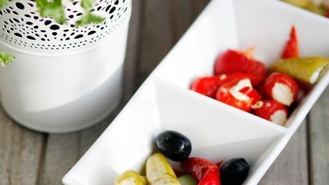 Eine längliche weiße Schüssel füllt sich mit bunten Antipasti, während frische Petersilie im Hintergrund steht, ideal zum Würzen für Salat.