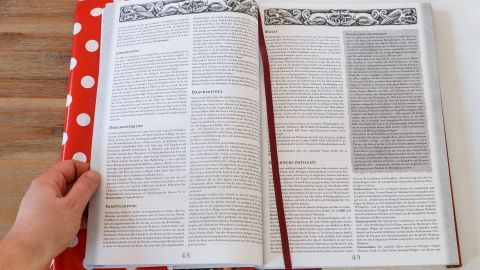 Eine Hand hält ein aufgeschlagenes Buch, schön geschützt durch eine bunte Hülle aus Wachstischdeckenstoff, auf einer Holzoberfläche.