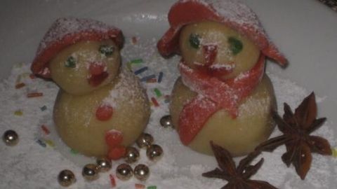 Zwei Marzipan-Schneemänner mit bunten Hüten und Schals auf einem Teller. Sie sind mit Puderzucker bestäubt, umgeben von Streuseln und Sternanis.