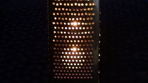 Eine alte Küchenreibe leuchtet als Windlicht und schafft eine gemütliche Atmosphäre im dunklen Raum.