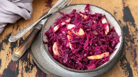 Ein bunter, marinierten Rotkohl-Salat mit Apfelstücken liegt in einer Schüssel auf einem rustikalen Holztisch.