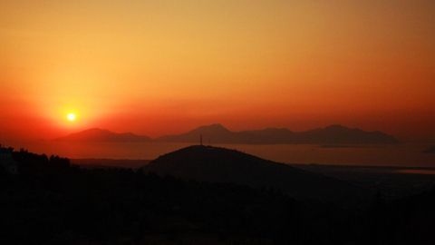 Ein Sonnenuntergang strahlt orange und rot über Hügel und Wasser, während die Silhouetten der Berge im Hintergrund sichtbar sind.