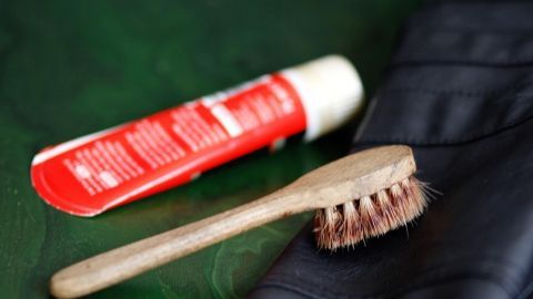 Eine abgenutzte schwarze Lederjacke liegt auf einer Oberfläche, während eine Bürste und rote Pflegecreme daneben für die Auffrischung bereitstehen.