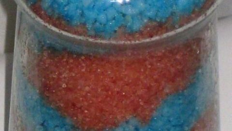 Ein Glasbehälter zeigt geschichtetes Badesalz mit abwechselnden Schichten aus blauem und rotem Salz auf einem weißen Hintergrund.