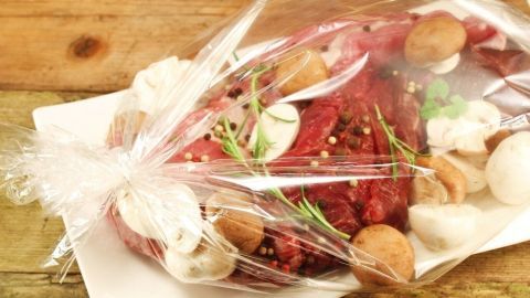 Ein Schweinebraten im Bratschlauch liegt auf einem weißen Teller, umgeben von frischen Kräutern und Pilzen.