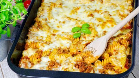Überbackene Nudeln Bolognese mit geschmolzenem Käse, serviert in einer Auflaufform, umrandet von frischen Kräutern.