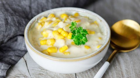 Eine Schüssel Maiscremesuppe steht auf einem rustikalen Holztisch, garniert mit frischer Petersilie und umgeben von einem goldenen Löffel.