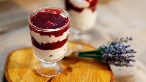 Ein Dessertglas mit Schichten aus cremigem Weiß und roter Fruchtsoße steht auf einem Holzbrett, umgeben von Blumen.