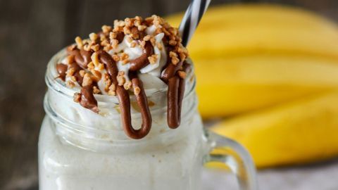 Ein cremiger Bananensplit-Milchshake in einem Glas mit Sahne, Schokoladensoße und Nüssen, umgeben von frischen Bananen.