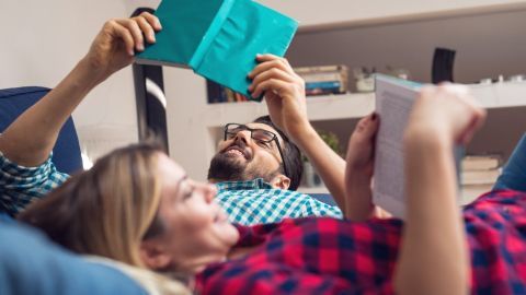 Ein Paar liest gemütlich auf einem Sofa. Beide halten Bücher in entspannter Atmosphäre, umgeben von Regalen im Hintergrund.