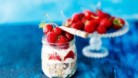 Ein Glas Bircher Müsli mit frischen Erdbeeren steht auf einem blauen Untergrund, während im Hintergrund eine Schale mit weiteren Erdbeeren sichtbar ist.