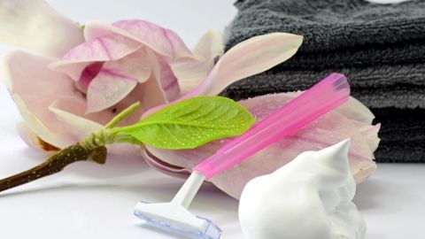 Wir zeigen dir, wie du Rasierpickel loswerden kannst! Ein Rasierer liegt auf einem Handtuch, umgeben von einer Blüte und einer Schaummasse, ideal für eine pickelfreie Bikinizone.