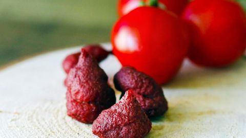 Tomatenmark ist günstiger in großen 850-ml-Dosen. Du kannst es in kleinen Portionen einfrieren und hast es somit beim Kochen immer griffbereit. Dunkles Tomatenmark liegt auf einem Holzbrett, während frische, rote Tomaten im Hintergrund stehen.