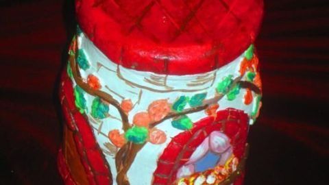 Ein bemaltes Schraubglas zeigt ein rot-goldenes Dach und Wände mit Blumenmustern. Es steht auf einem dunklen Untergrund.