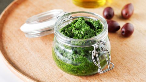 Ein Glas mit grünem Pesto steht auf einem Holzbrett, umgeben von Olivenöl und einigen Oliven.