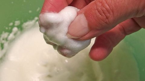 Eine Hand hält einen weißen, schaumigen Klumpen, während sie sich über einer grünen Schüssel mit Eiweißmaske beugt.
