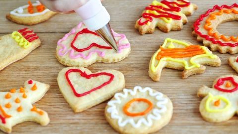Eine Hand benutzt eine Spritztüte, um Zuckerschrift auf ausgebackene Plätzchen mit bunten Verzierungen anzubringen.