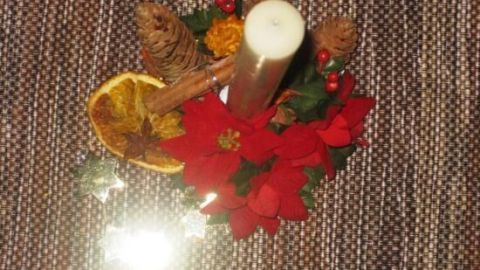 Ein Weihnachtsgesteck aus roten Blüten, einer Kerze, getrockneten Orangenscheiben und Tannenzapfen auf einem Tisch.