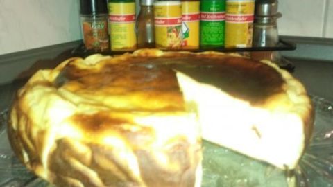 Käsekuchen ohne Boden sitzt auf einem Glasteller in der Küche, mit einer herausgeschnittenen Scheibe. Im Hintergrund Gewürze.