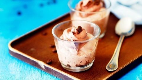 Zwei Gläser mit Latte-Macchiato-Creme stehen auf einem Tablett, umgeben von einem blauen Hintergrund und einer Kaffeekanne.