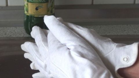 Weiße Öl-Handschuhe liegen faltenfrei auf einer Arbeitsfläche, während zwei Hände sie sanft übereinanderlegen, wobei ein Getränk im Hintergrund sichtbar ist.